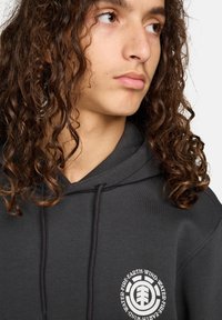 Sudadera negra con cordones, que presenta un logotipo circular blanco en el lado izquierdo del pecho con texto. Textura de tela suave y corte casual.