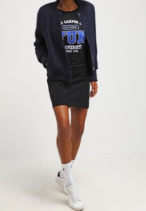 Person trägt eine marineblaue Bomberjacke über einem schwarzen T-Shirt mit Grafik, schwarzen Rock, weißen Socken und weißen Sneakers und geht nach vorne.