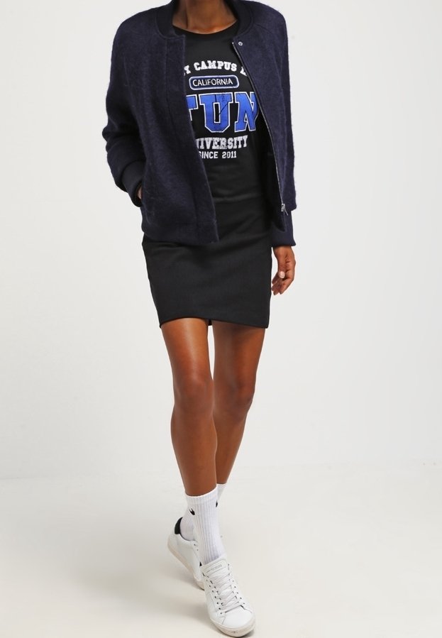 Personne portant une veste bomber bleu marine sur un t-shirt graphique noir, une jupe noire, des chaussettes blanches et des baskets blanches marchant vers l'avant.