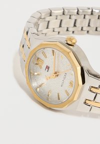 Ασημένιο και χρυσό μεταλλικό ρολόι χειρός με οκταγωνικό πλαίσιο, χρυσούς δείκτες ώρας και την επωνυμία "Hilfiger" σε υφασμένο λευκό καντράν.