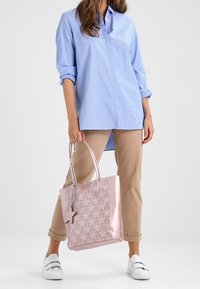 Ljusrosa tote-väska med ett mönstrat överdrag och en stjärnformad detaljer. Bärs med en blårandig skjorta och beiga byxor, vilket visar en avslappnad passform.