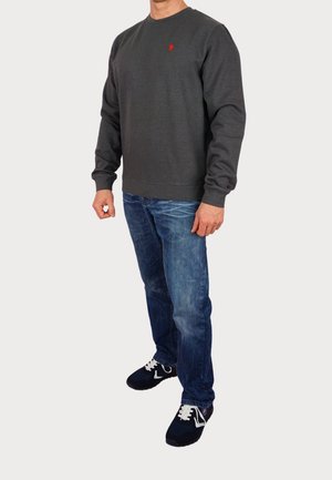 Sudadera gris con escote redondo y logo rojo, combinada con jeans de mezclilla azul y zapatillas negras con cordones blancos. Ajuste informal.