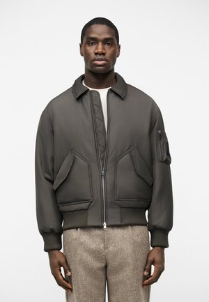 Modèle masculin portant une veste bomber olive foncé avec des poches avant et une poche zippée sur la manche, associée à un pantalon en laine beige sur fond blanc.