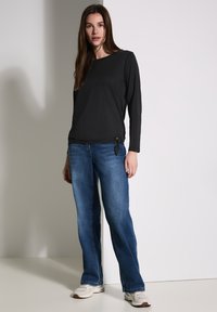 Maglietta nera a maniche lunghe con dettaglio nodo in vita, abbinata a jeans dritti azzurri chiari e sneakers bianche su uno sfondo neutro.