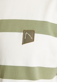 Dryžuota medvilninė marškinė su pakaitomis baltomis ir alyvuogių žaliais horizontaliais dryžiais, su tekstūriniu trikampiu logotipu priekyje.