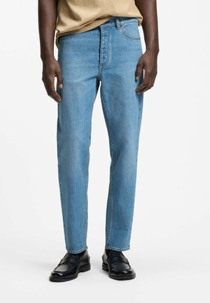 Mand iført lyseblå jeans med lige ben, beige top og sorte læderloafers stående mod en ensartet hvid baggrund.