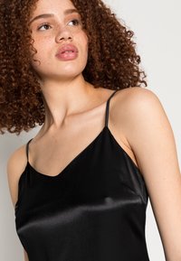 Camisola de cetim preta com alças finas, apresentando uma textura suave e um decote em V profundo, destacando o seu design elegante e minimalista.