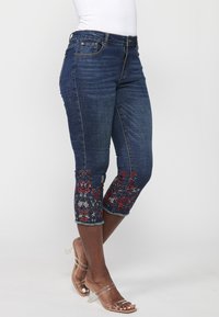 Denim capri jeans in dark blue with floral embroidery in red and blue at the cuffs. Features a classic fit and slight stretch. Clear-heeled sandals.