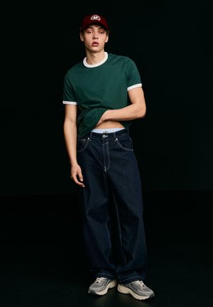 Fiatal férfi sötétzöld pólóban, sötét, bőszárú farmerral, szürke tornacipőben és piros baseballsapkában, fekete háttér előtt.