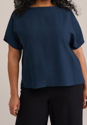 Kvinde iført en løs mørkeblå kortærmet top og sorte bukser, stående mod en ensfarvet neutral baggrund.