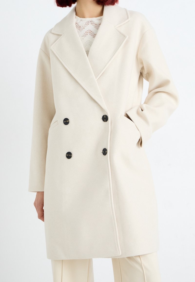 Femme portant un manteau en laine crème à double boutonnage avec des boutons noirs et des poches latérales, associé à un pantalon assorti et un top en dentelle.