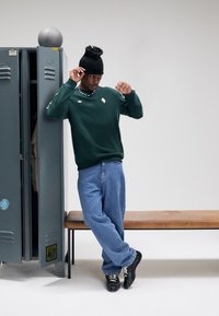 Jovem com sweat verde, jeans azuis e gorro preto posando junto a cacifos cinzentos abertos e um banco de madeira numa sala branca.