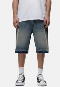 Shorts in denim blu chiaro con una finitura sbiadita, orlo grezzo e un accento blu scuro a contrasto in vita, indossati con sneakers bianche e nere.
