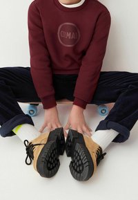 Bordeaux huppari, jossa on pyöreä logo keskellä, yhdistetty tummiin samettilapasaumoihin ja vaaleisiin kenkiin, joissa on mustat kumipohjat.