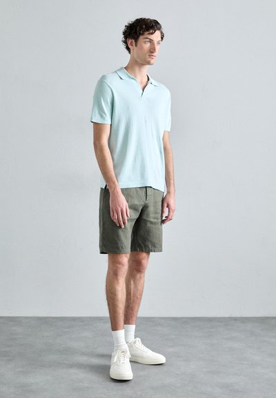Polo shirt en coton bleu clair avec un col plat, associé à un short vert olive. Des baskets blanches et des chaussettes blanches de longueur mi-mollet complètent la tenue.