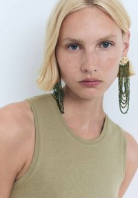 Beaded earrings featuring green strands, embellished with gold accents, paired with a fitted olive tank top. Smooth fabric texture.