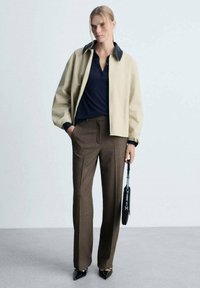 Veste bomber beige avec col noir, polo bleu marine, pantalon brun ajusté, escarpins noirs pointus, tenant un petit sac à main noir.
