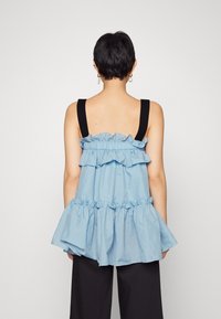 Top a peplo azzurro con volant, spalline nere, caratterizzato da un design a strati e un tessuto testurizzato. Indossato sopra pantaloni neri.
