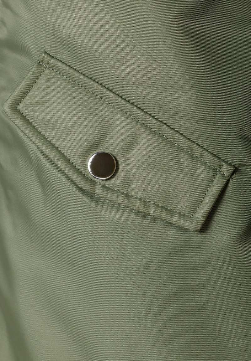 Tela verde oliva con una textura suave presenta una solapa de bolsillo diagonal asegurada por un botón a presión plateado.