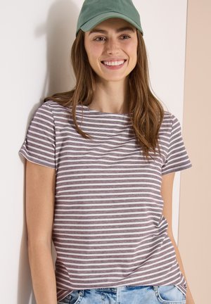 Jeune femme souriante, portant une casquette verte, un t-shirt rayé gris et blanc à manches courtes et un jean bleu clair, debout devant un mur bicolore.