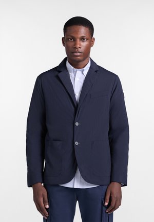 Muž v tmavomodrom saku cez svetlomodrú pruhovanú košeľu, stojaci pred jednoduchým bielym pozadím.