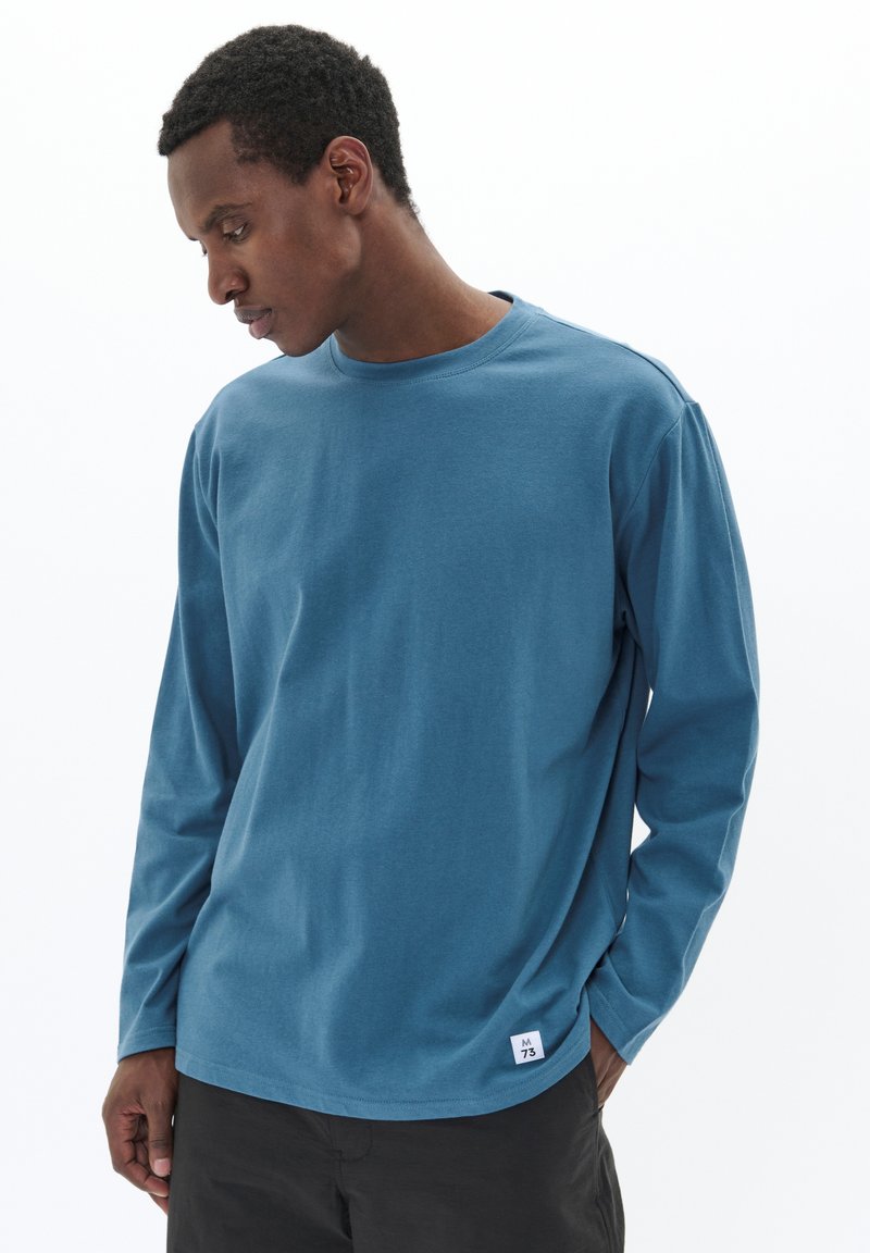 Langärmliges blaues Baumwoll-T-Shirt mit Rundhalsausschnitt, subtiler Textur und einem kleinen weißen Etikett in der unteren linken Ecke.