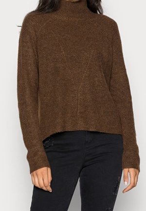 Femme portant un pull en maille marron à col montant et un jean noir déchiré, debout devant un fond uni.