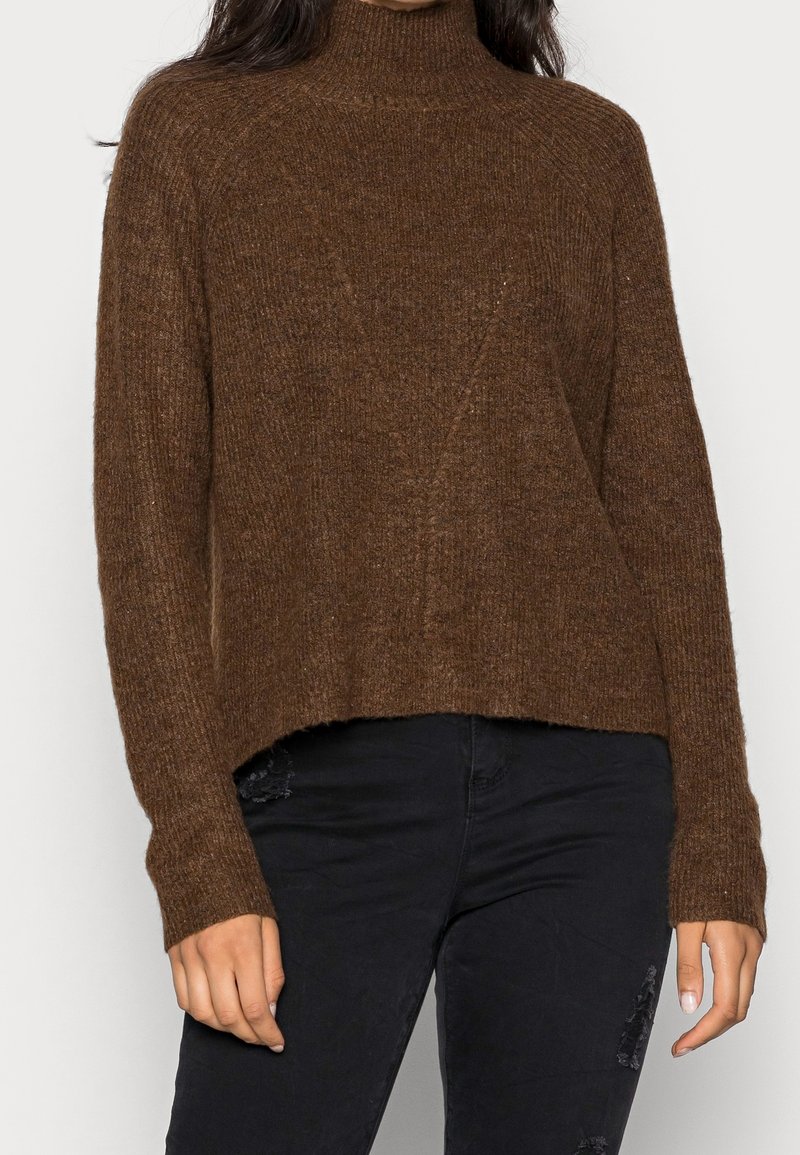 Femme portant un pull en maille marron à col montant et un jean noir déchiré, debout devant un fond uni.