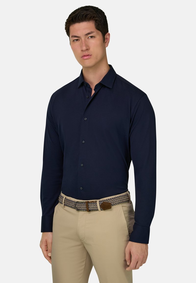 Jeune homme portant une chemise boutonnée bleu marine, un pantalon beige et une ceinture tressée, debout devant un fond clair uni.