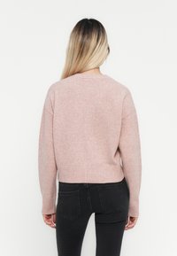 Rosa stickad tröja med en kortad design, ribbade ärmslut och slut. Modellen bär svarta jeans och visar tröjans avslappnade silhuett bakifrån.