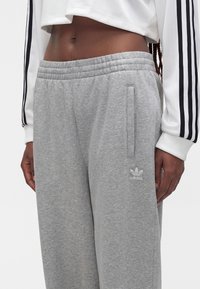 Suurplaan inimesest, kes kannab halli Adidas dressipükse ja valget pikkade varrukatega pluusi mustade triipudega varrukaotstel.