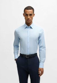 Hellblaues Langarmhemd mit Button-Down-Kragen, glatter Textur und weißen Knöpfen, kombiniert mit dunklen gemusterten Hosen und einem schwarzen Gürtel.