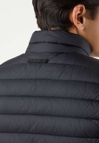 Uomo che indossa una giacca trapuntata nera con colletto alto, mostrato di schiena e di lato con particolare attenzione alla zona della spalla e del collo.