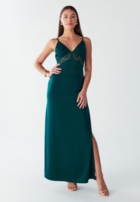 Vestito maxi in satin verde mare con inserto in pizzo in vita, scollatura a V profonda, spalline sottili e uno spacco laterale. Texture morbida e vestibilità elegante.
