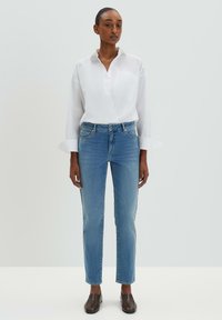 Chemise blanche à manches longues avec une poche, jean slim bleu, et chaussures brunes à enfiler. Tissu lisse, design simple, portée debout contre un fond clair.