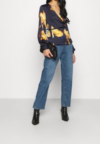 Marinebluse mit gelbem Schmetterlingsdruck, Wickeldesign, lange Ärmel. Kombiniert mit schmalen Jeans und schwarzen Ankle Boots, hält eine schwarze Clutch.