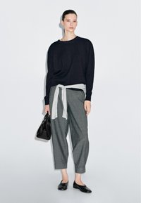 Maglione nero a maglia indossato sopra una camicia grigia, pantaloni grigi legati con una cintura grigia chiara, borsa nera e scarpe nere, su uno sfondo semplice.