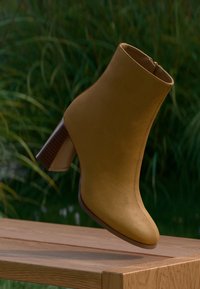 Gul skinn ankelstøvel med rund tå, brun trekloss hæl og glidelås på siden, satt mot en uskarp bakgrunn.