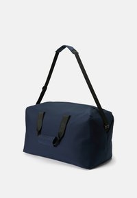 Marineblå duffelbag laget av slitesterkt materiale, med to svarte håndtak og en justerbar skulderrem for variert bærealternativer.