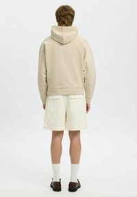 Felpa beige con cappuccio, texture morbida e polsini a coste, abbinata a pantaloni corti avorio con tasche posteriori. Design semplice, colori neutri.
