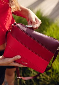 Červená a vínová crossbody taška z texturované kůže, se zipovým uzávěrem, viditelným prošíváním a zlatým logem jako akcentem. Obsahuje nastavitelný popruh.