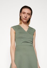 Vestido verde, ajustado, com decote em V e mangas curtas, apresentando uma textura suave e um design envolvente na cintura. Silhueta simples e elegante.