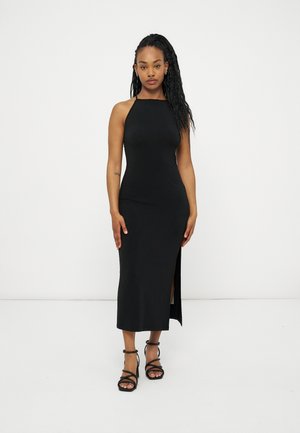 Γυναίκα όρθια, φοράει μαύρο μίντι φόρεμα χωρίς μανίκια με πλευρική σκισμή και μαύρα ψηλοτάκουνα πέδιλα με λουράκια, σε απλό λευκό φόντο.