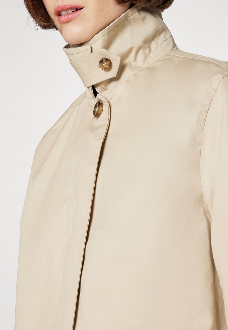 Chaqueta beige con un alto cuello, dos botones redondos y una textura suave, que enfatiza el diseño minimalista y un estilo funcional.