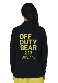Schwarzes Langarmshirt aus strukturiertem Stoff. Gelber Grafiktext auf der Rückseite mit der Aufschrift "OFF DUTY GEAR 223" und einer Stadtlandschaftsgrafik.