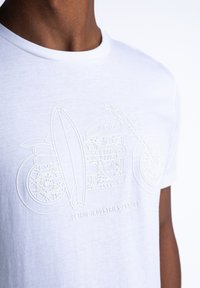 Biała koszulka z wytłoczonym wzorem przedstawiającym motocykl i deskę surfingową, z napisem "Petrol Industries Est. 89" poniżej.