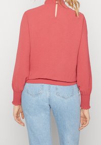 Koralli, pitkähihainen toppi, jossa on rypytetty kangas pääntiellä ja hihojen päällä, selässä pieni avainreikäyksityiskohta; yhdistettynä vaaleansiinisiin farkkuihin.