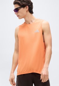 Νεαρός άνδρας φορώντας πορτοκαλί αμάνικη μπλούζα Adidas και μαύρο σορτς, κοιτάζει προς τα αριστερά του, με αθλητικά γυαλιά ηλίου.