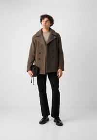 Brun ull peacoat med overdimensjonert krage, dobbelknappede knapper, svarte detaljer, parret med en beige topp og svarte bukser. Svarte vesker i hånden.