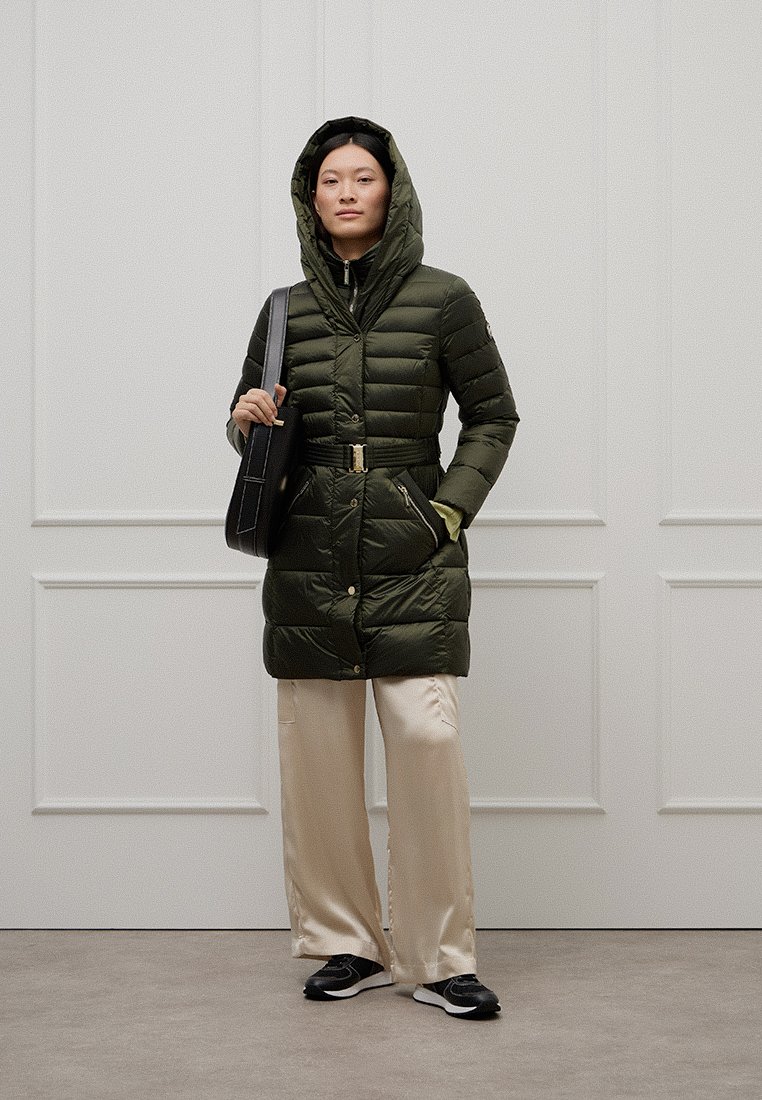 Frau in einem olivgrünen, taillierten Daunenmantel mit Kapuze, hellbeigen, weiten Hosen, schwarzen Turnschuhen und einer schwarzen Umhängetasche.